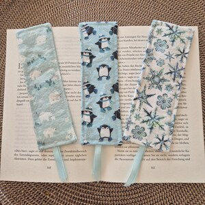 Könnte beinhalten: Drei Stoff-Lesezeichen mit hellblauen Bändern liegen auf einem Buch. Eines zeigt Eisbären, ein anderes Pinguine und das dritte Schneeflocken, alle auf hellblauem Hintergrund. Die rechteckigen Lesezeichen haben einen genähten Rand.