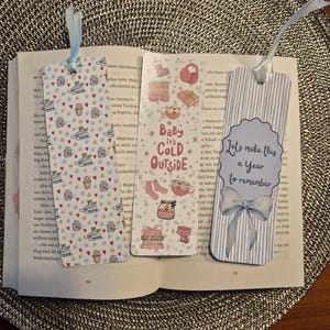 Könnte beinhalten: Drei dekorative Lesezeichen mit Schleifen liegen auf einem offenen Buch. Eines zeigt Eislaufschuhe, Herzen und Kaffeetassen. Ein anderes sagt "Baby it's Cold Outside" mit Winter-Icons. Das dritte sagt "Let's make this a year to remember".