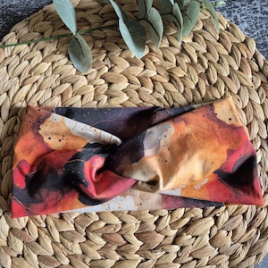 Könnte beinhalten: Ein farbenfrohes Stirnband mit einem gedrehten Knoten. Der Stoff zeigt ein lebendiges abstraktes Muster in Rot-, Orange-, Schwarz- und Cremetönen. Das Stirnband liegt auf einem gewebten, runden Platzdeckchen.