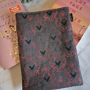 Könnte beinhalten: Eine rechteckige Buchhülle aus Stoff mit dunkelgrauem Hintergrund, einem sich wiederholenden roten Damastmuster und kleinen schwarzen Fledermaussilhouetten. Die Hülle hat ein kleines schwarzes Stoffetikett an der Seite und sichtbare Nähte.