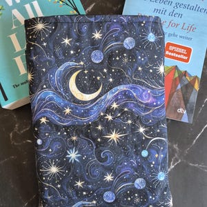 Könnte beinhalten: Ein dunkelblauer Stoff-Buchumschlag mit einem Himmelsdesign mit Mond, Sternen und Wirbelmustern. Der Umschlag ist um ein Buch gewickelt, dessen Titel teilweise sichtbar ist. Das Design umfasst Blau-, Weiß- und Goldtöne.