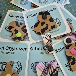 May include: A collection of cable organisers featuring various shapes and patterns, including heart-shaped leopard print, flower-shaped tan, and plaid designs. Each organiser is attached to a light blue card labelled Kabel Organizer Handmade.