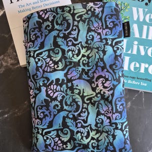 Könnte beinhalten: Eine rechteckige Buchhülle aus Stoff mit schwarzem Hintergrund und einem sich wiederholenden Muster aus Katzen in Blau-, Lila- und Türkistönen, durchsetzt mit wirbelnden Blumenmotiven. An der Seitenkante befindet sich ein kleines schwarzes Stoffetikett.
