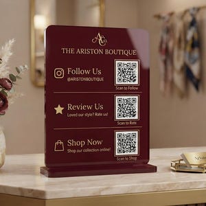 May include: A rectangular dark red acrylic sign featuring the gold text THE ARISTON BOUTIQUE, three QR codes, and icons for Instagram, a star, and a shopping bag. The sign includes the text Follow Us, Review Us, and Shop Now with scan prompts.