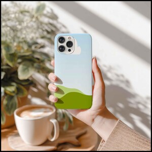 May include: A light blue and white phone case with a landscape design featuring green hills and a white cloud. The case is held in a hand, with a coffee cup and plant in the background.