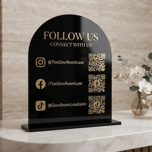 May include: A black arched sign with gold text and icons featuring the words FOLLOW US and CONNECT WITH US. It displays Instagram, Facebook, and TikTok social media handles and corresponding QR codes for TheGlowRoomLuxe and GlowRoomLuxeSalon.