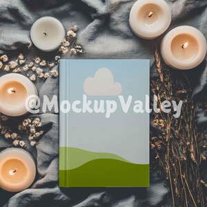 May include: A blue and green journal with a white cloud and a green hill design. The journal is surrounded by white candles and dried flowers on a gray fabric background.