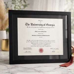 May include: A black framed diploma from The University of Georgia, with a red tassel. The diploma certifies a Bachelor of Business Administration degree. The frame is rectangular and sits on a white surface.