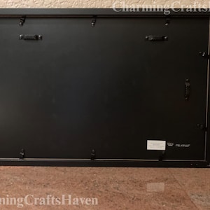 May include: Black rectangular picture frame with a black backing. The frame has metal hardware for hanging and securing the backing. The frame is on a brown surface. The text "Charming Crafts Haven" is at the bottom.