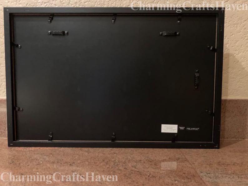 May include: Black rectangular picture frame with a black backing. The frame has metal hardware for hanging and securing the backing. The frame is on a brown surface. The text "Charming Crafts Haven" is at the bottom.