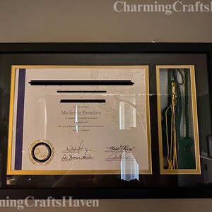 May include: A framed diploma and graduation tassel. The diploma is in a gold frame with a black border, and the tassel is in a separate compartment. The diploma has a seal and signatures. The frame is hanging on a wall.