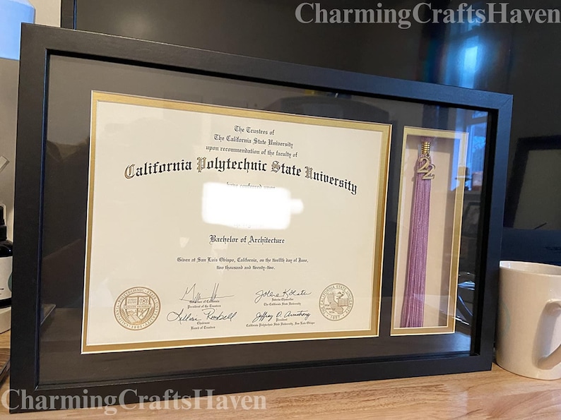 May include: Black framed diploma from California Polytechnic State University, Bachelor of Architecture, with a gold border. A separate compartment holds a purple tassel with a gold "22" charm. The frame is on a wooden surface.