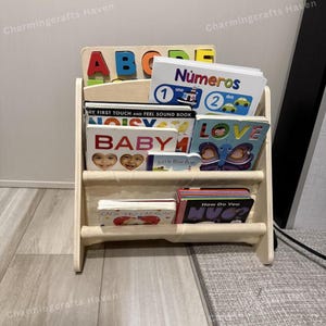 May include: A wooden bookshelf filled with children's books and a wooden ABC puzzle. The books have colorful covers and titles like "Baby" and "Love." The bookshelf has a light-colored fabric sling for holding books.