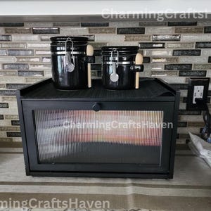 May include: Black ceramic canisters with wooden spoons on a black countertop with a black bread box with a glass door. The countertop is tiled with a mosaic of gray and brown tiles.