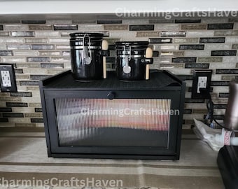 May include: Black ceramic canisters with wooden spoons on a black countertop with a black bread box with a glass door. The countertop is tiled with a mosaic of gray and brown tiles.