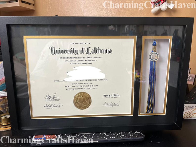 May include: A framed University of California diploma with a gold seal and a blue and gold tassel. The diploma is in a black frame with a gold border. The text on the diploma reads "University of California".