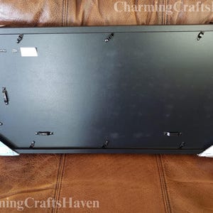 May include: A rectangular black picture frame with hanging hardware on the back. The frame is protected by white corner protectors. The frame is resting on a brown leather surface.