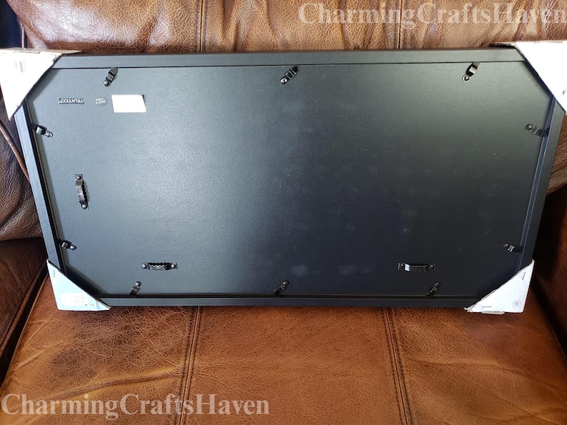 May include: A rectangular black picture frame with hanging hardware on the back. The frame is protected by white corner protectors. The frame is resting on a brown leather surface.