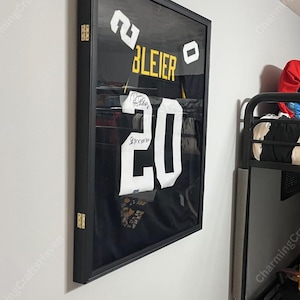 May include: Black framed jersey with the name "BLEIER" and the number "20" in white and gold. The jersey is displayed on a white wall. The frame has gold hinges.