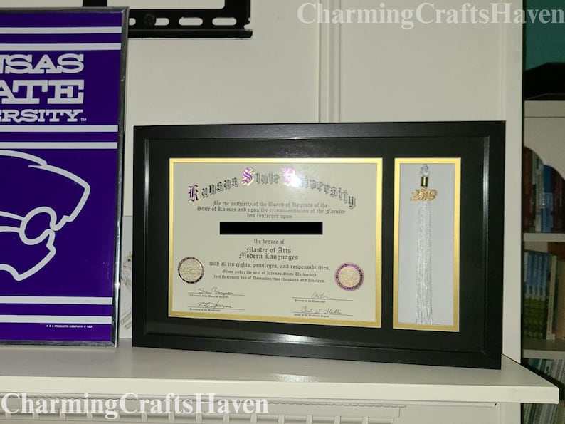 May include: A framed diploma from Kansas State University, with a black frame and gold accents. The diploma is for a Master of Arts in Modern Languages. A tassel with a 2019 charm is displayed next to the diploma.