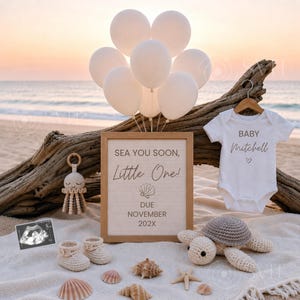 Op de afbeelding: Een witte babyromper met de tekst BABY Mitchell en een hartje, een letterbord met de tekst SEA YOU SOON, LITTLE One! DUE NOVEMBER 202X, een gehaakte kwal, een gehaakte schildpad, babyslofjes, een echo en witte ballonnen op een strand.