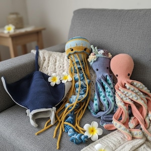 May include: A collection of handmade crochet sea creatures, including a blue manta ray, a jellyfish with yellow and blue accents, a purple octopus, and a peach octopus. The toys are arranged on a gray couch with decorative flowers.