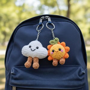 May include: Two crochet keychains hang from a blue backpack zipper. One is a white cloud with brown legs, the other is a smiling orange sun with green leaves. Both have black eyes and are attached to silver keyrings.