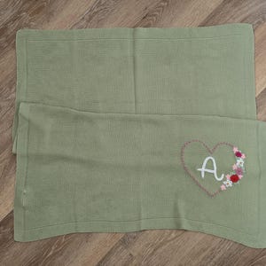May include: A sage green knitted blanket featuring a rectangular shape with a border. The blanket is embroidered with a large heart outline in a darker thread, containing a white letter A and a cluster of small colourful floral accents.