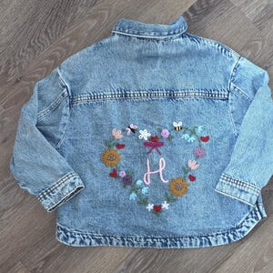 May include: A light blue denim jacket featuring a back panel embroidered with a heart-shaped wreath of colourful flowers, leaves, a small bee, and a pink letter H in the centre. The jacket has a classic collar, long sleeves, and button cuffs.