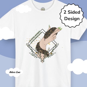 Puede incluir: Camiseta blanca con un gráfico de una mano sosteniendo una serpiente marrón y negra, con el texto "In every moment you're always there." El diseño está enmarcado por un diamante negro y hojas verdes. El texto "2 Sided Design" está en un círculo negro.