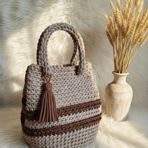 May include: A handmade crochet tote bag in beige and brown. The bag features a braided handle, a decorative tassel, and two horizontal brown stripes. The bag is sitting on a white, fluffy surface, with a vase of wheat stalks in the background.