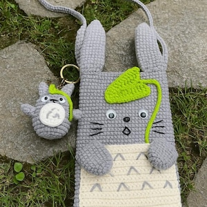 May include: A handmade grey crochet bag and matching keyring. The bag features a cartoon character design with large ears, a leaf, and a cream-coloured pocket. The keyring is a smaller version of the bag's character.