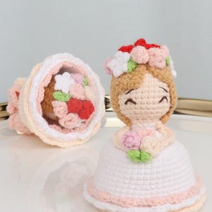Op de afbeelding: Gehaakte pop in een witte jurk met een bloemenkrans en een boeket. De pop heeft een lachend gezicht en is gemaakt met lichtbruin, wit, roze en rood garen. Een tweede gehaakt boeket is ook zichtbaar.