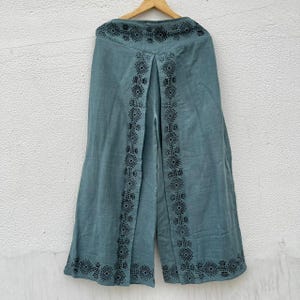 May include: Teal-colored wide-leg pants with black geometric embroidery along the sides and hem. The pants are hanging on a wooden hanger against a white wall. The embroidery features diamond and square patterns.