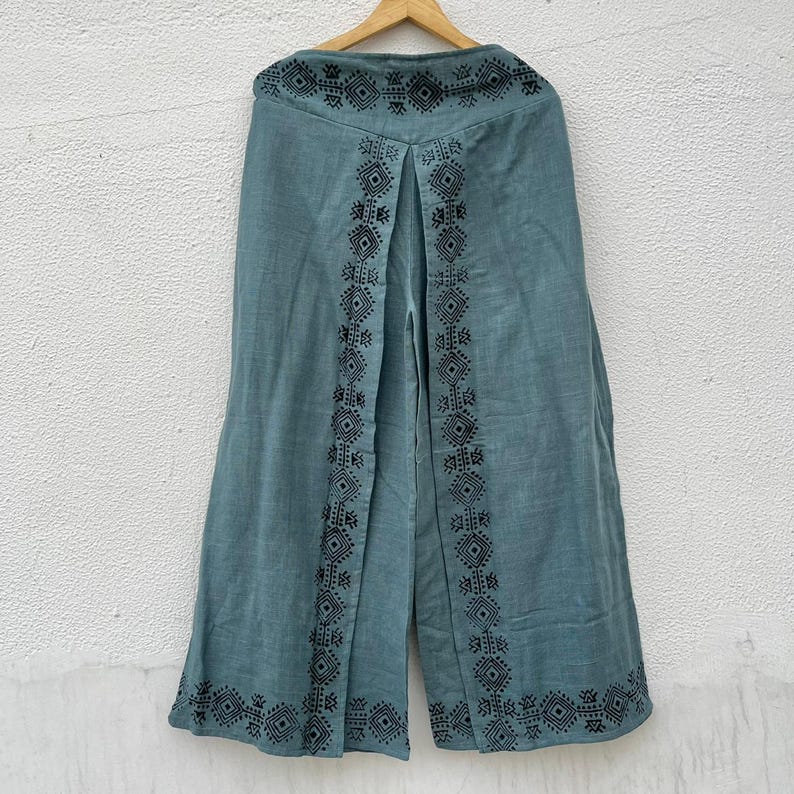 May include: Teal-colored wide-leg pants with black geometric embroidery along the sides and hem. The pants are hanging on a wooden hanger against a white wall. The embroidery features diamond and square patterns.