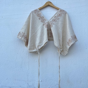 May include: A cream-colored cropped top featuring a deep V-neckline and wide, short sleeves. The garment is decorated with brown geometric patterns along the neckline and sleeve edges, and includes two long, thin drawstrings at the bottom hem.