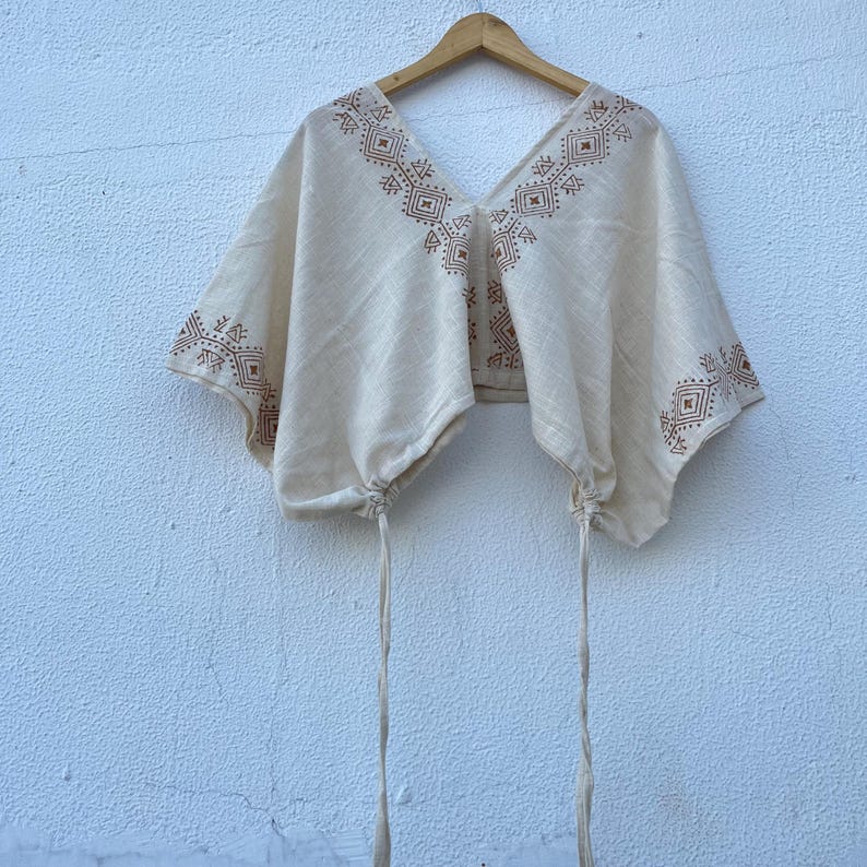 May include: A cream-colored cropped top featuring a deep V-neckline and wide, short sleeves. The garment is decorated with brown geometric patterns along the neckline and sleeve edges, and includes two long, thin drawstrings at the bottom hem.
