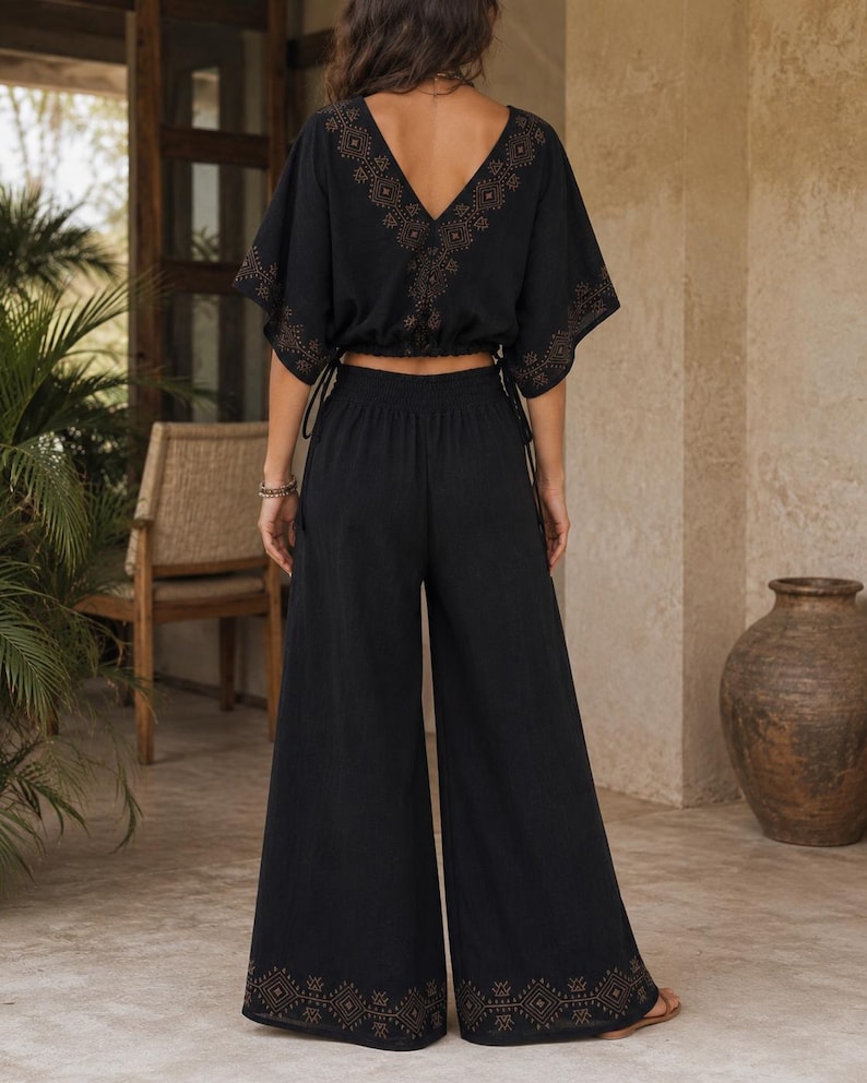 May include: Black two-piece outfit featuring a V-neck top with short sleeves and wide-leg pants. The top and pants have decorative patterns in a contrasting color. The outfit is made of a lightweight fabric, suitable for warm weather.