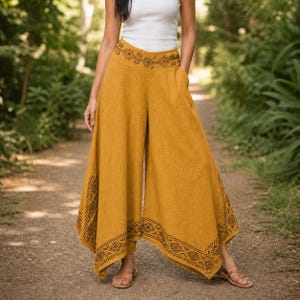 May include: Mustard-colored palazzo pants with a wide leg design. The pants feature an embroidered pattern along the waistband and hem. The model is wearing a white tank top and sandals. The pants are made of a lightweight fabric.