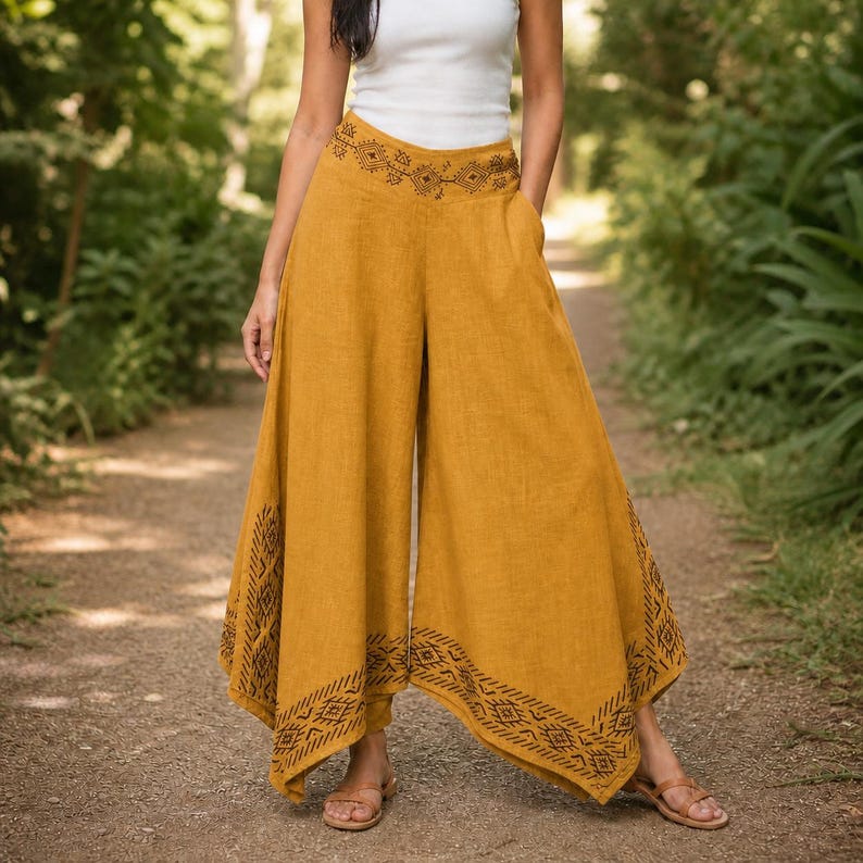 May include: Mustard-colored palazzo pants with a wide leg design. The pants feature an embroidered pattern along the waistband and hem. The model is wearing a white tank top and sandals. The pants are made of a lightweight fabric.
