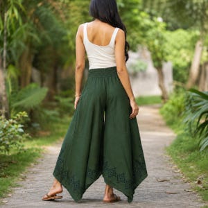 May include: A woman wearing a white tank top and dark green palazzo pants. The wide-leg pants have an elastic waistband and a geometric pattern near the hem. The pants are made of a flowing fabric, suitable for warm weather.