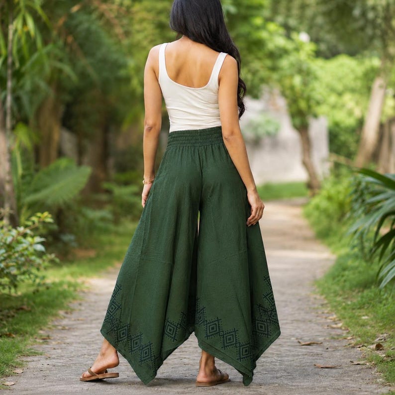 May include: A woman wearing a white tank top and dark green palazzo pants. The wide-leg pants have an elastic waistband and a geometric pattern near the hem. The pants are made of a flowing fabric, suitable for warm weather.