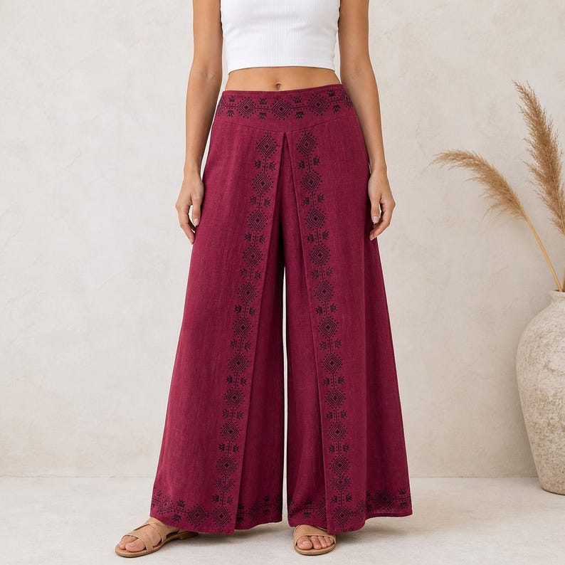 May include: Wide-leg burgundy pants with black geometric embroidery down the front. The pants have a high waist and a split front design. The model is wearing a white crop top and sandals.