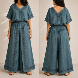 May include: A two-piece teal outfit featuring a V-neck top and wide-leg pants. The top has short sleeves and an elastic waist. The pants have a matching elastic waist and a decorative embroidered design down the front. The outfit is made of a soft, flowing fabric.