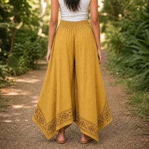 May include: Mustard yellow palazzo pants with a wide leg and elastic waistband. The pants feature a geometric pattern at the hem. The model is wearing a white tank top and sandals. The pants are made of a lightweight fabric.