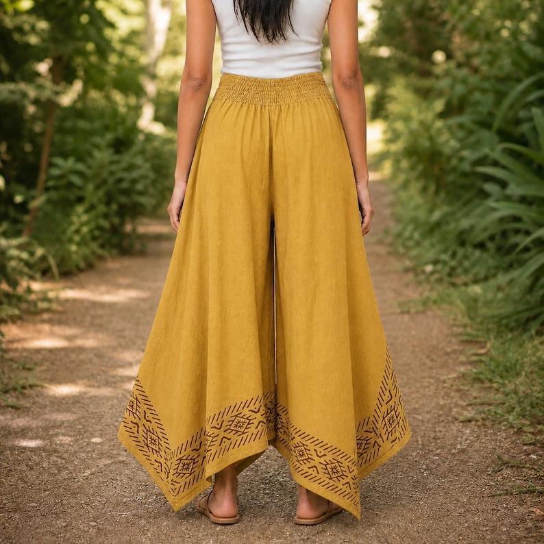 May include: Mustard yellow palazzo pants with a wide leg and elastic waistband. The pants feature a geometric pattern at the hem. The model is wearing a white tank top and sandals. The pants are made of a lightweight fabric.
