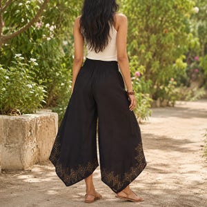 May include: Black palazzo pants with a wide leg and an elastic waistband. The pants feature a geometric pattern in a gold color along the bottom hem. The model is wearing a white tank top and sandals.