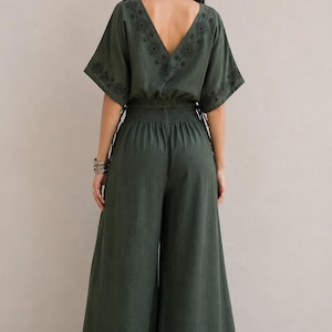 May include: A dark green jumpsuit with a V-neckline and wide-leg pants. The jumpsuit features embroidered detailing along the neckline, sleeves, and pant hems. The waist is cinched with an elastic band. The model is wearing sandals.