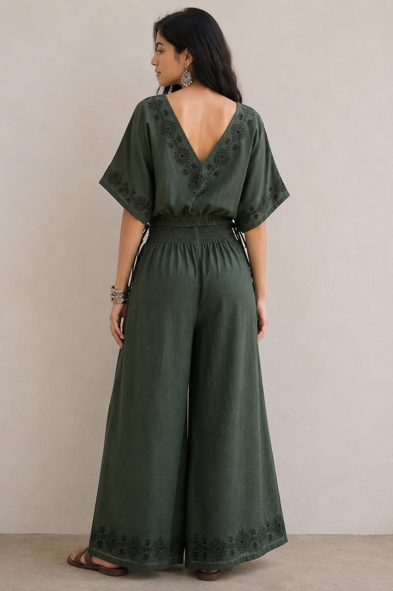 May include: A dark green jumpsuit with a V-neckline and wide-leg pants. The jumpsuit features embroidered detailing along the neckline, sleeves, and pant hems. The waist is cinched with an elastic band. The model is wearing sandals.
