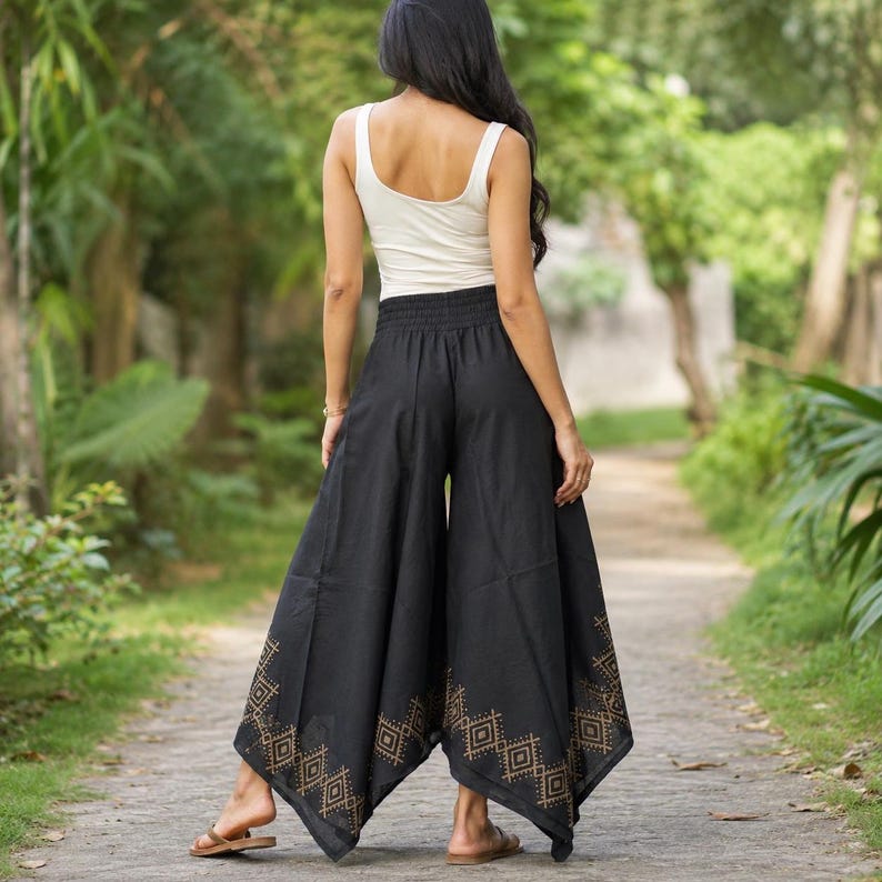 May include: Black palazzo pants with a wide leg and an elastic waistband. The pants feature a geometric pattern in a gold color at the hem. The person in the image is wearing a white tank top and sandals. The background is a pathway surrounded by greenery.